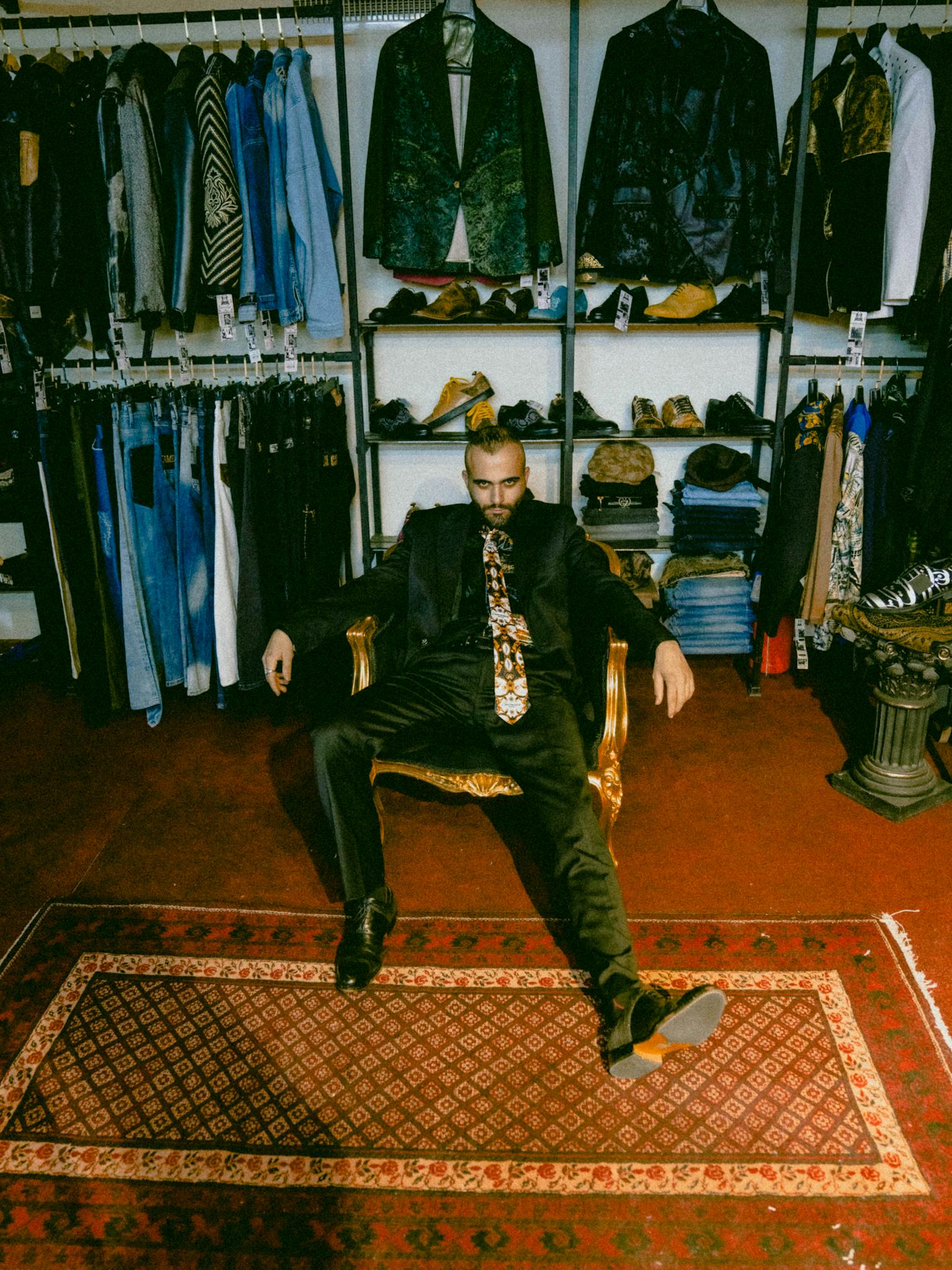 Man in a stylish clothing store, seated on a chair with arms outstretched, amid tailored suits and shoes.